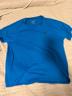 Blå tränings-tshirt från Under Armour - Snygg blå tränings-tshirt från Under Armour med rund hals och korta ärmar. Tillverkad i ett lätt och snabbtorkande funktionsmaterial, perfekt för gymmet eller löprundan. Diskret logga på bröstet.