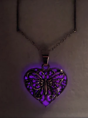 Halsband med hjärthänge och fjäril - Lyser i mörker  - Unikt halsband med ett hjärtformat hänge i silverfärgad metall och ett detaljerat fjärilsmotiv. Hänget har en rosa bakgrund som lyser lila i mörker, vilket ger en cool glow-effekt. Vanligt dagsljus aktiverar effekten i hänget, solljus fungerar bäst, vänd på hänget en kort stund i dagljus eller solljus för att få bästa effekt i mörker. Kedjan är tunn och justerbar i rostfritt stål. Tål vatten utan att missfärgas. Perfekt för dig som gillar statement-smycken med magisk känsla.