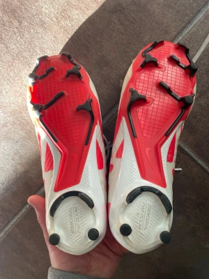 Nike vita och röda fotbollsskor - Säljer ett par Nike fotbollsskor med vita och röda detaljer samt svart swoosh på ovansidan. Skorna har snörning och en robust sula med dobbar för bra grepp på planen. Materialet är syntetiskt och designen är modern och sportig. Perfekta för dig som vill ha stil och funktion på fotbollsplanen.