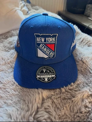 Blå New York Rangers keps - Snygg blå keps från Mitchell & Ness med New York Rangers-logga broderad framtill. Kepsen har en klassisk form och sticker ut med detaljer på sidorna och en sticker på skärmen. Perfekt för dig som gillar sport och streetstyle.