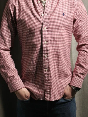 Rutig röd skjorta från Ralph Lauren - Klassisk röd och vit rutig skjorta från Ralph Lauren med blå logga broderad på bröstet. Skjortan har lång ärm, knappar framtill och krage. Tillverkad i bomull för en bekväm känsla och snygg passform.