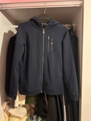 Mörkblå hoodie från Sail Racing - Mörkblå hoodie från Sail Racing med dragkedja framtill och en extra ficka med dragkedja på bröstet. Klassisk huva och diskret logga på bröstet. Tillverkad i mjukt material som känns skönt mot huden. Perfekt för en avslappnad och sportig stil.