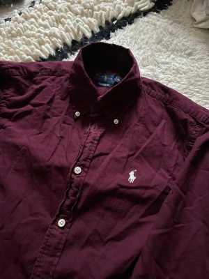 Vinröd skjorta från Ralph Lauren - Vinröd långärmad skjorta från Ralph Lauren med klassisk button-down krage och vit broderad logga på bröstet. Skjortan har vita knappar och är tillverkad i bomull. Perfekt för en stilren look.