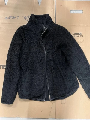 Svart teddyjacka med dragkedja - Mysig svart teddyjacka med hög krage och hel dragkedja framtill. Jackan har två sidofickor och är tillverkad i fluffigt teddy-material som håller dig varm. Perfekt för kyliga dagar och enkel att matcha med olika outfits. Använt skick men hel. 