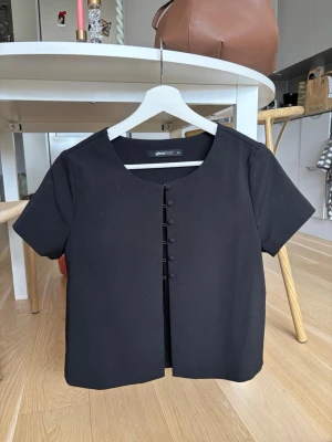 Svart kortärmad blus från Gina Tricot - Stilren svart blus från Gina Tricot med rund halsringning och klädda knappar framtill. Kortärmad modell i mjukt tyg som ger en clean och modern look. Perfekt att matcha med jeans eller kjol för en enkel men snygg outfit.