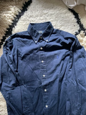 Mörkblå skjorta från Polo Ralph Lauren - Klassisk mörkblå skjorta från Polo Ralph Lauren med button-down krage och broderad logga på bröstet. Skjortan har långa ärmar, vita knappar och är tillverkad i mjuk bomull. Perfekt för en clean och stilren look.