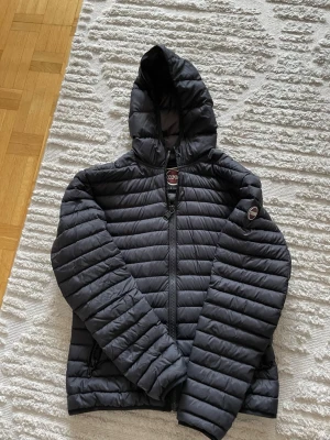 Svart dunjacka med huva från Colmar - Snygg svart dunjacka från Colmar med huva och dragkedja framtill. Jackan har två sidofickor med dragkedja och Colmar-logga på ärmen. Lätt vadderad och perfekt  året om. Klassisk quiltad design som håller värmen.