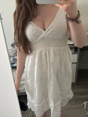 Vit kort klänning med broderi - Söt vit kort klänning med v-ringning och breda axelband. Klänningen har fina broderade blommönster och volangkant nertill. Tillverkad i lätt bomull, perfekt för varma dagar. Smockad midja för bekväm passform och luftig känsla.