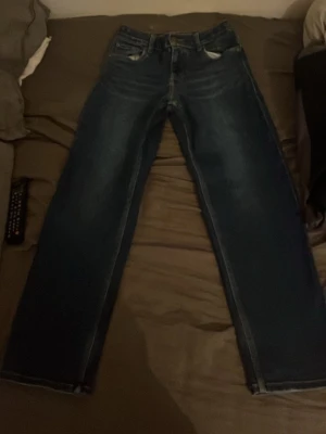 Mörkblå raka jeans från Levi's - Säljer ett par klassiska mörkblå jeans från Levi's med raka ben och femficksmodell. Jeansen har subtila slitningar framtill och bakfickor med den ikoniska Levi's-lappen. Tillverkade i slitstark denim, perfekta för en avslappnad och tidlös stil.