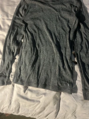 Mörkgrå långärmad ullunderställ tröja - En stilren mörkgrå långärmad ullunderställ tröja i mjukt och stretchigt material från märket craft. Perfekt för träning eller som baslager. Tröjan har en enkel design utan tryck och är lätt att matcha med andra plagg. I junior storlek