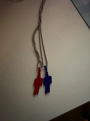 Halsband med röda och blå figurer - Unikt halsband med två hängsmycken i form av stiliserade figurer, en i rött och en i blått. Figurerna är tillverkade i plast med glittereffekt och hänger på en tunn silverfärgad kedja. Perfekt accessoar för dig som gillar färg och lekfull design. Billie eilish halsband