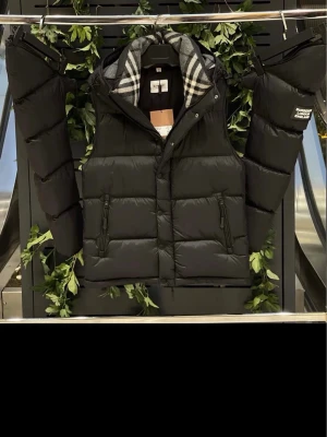 Svart dunväst från Burberry - Snygg svart dunväst från Burberry med avtagbara ärmar och klassiskt rutigt foder i huvan. Västen har två dragkedjefickor framtill och en diskret logotyp på ena ärmen. Perfekt för lager-på-lager och streetstyle.