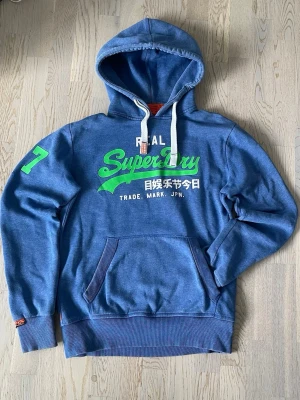 Blå Superdry hoodie med tryck - Blå hoodie från Superdry med stor grön och vit logga på bröstet och japansk text. Har en stor ficka framtill, vita dragsnören i huvan och en grön siffra 77 på ärmen. Mjuk bomullsblandning och ribbade muddar. Perfekt för en avslappnad streetwear-look. Lite nött under siffrorna. Sorry för upp och ner bilder. Mycket bra skick annars.