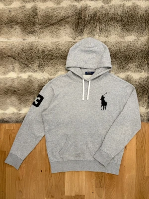 Ljusgrå Polo Ralph Lauren Hoodie, Big Pony - Storlek L - Hej! 👋 Säljer en riktigt snygg ljusgrå Polo Ralph Lauren hoodie i storlek L. Det här är "Big Pony"-modellen med den stora svarta loggan på bröstet och den ikoniska "3:an" broderad på höger ärm – en detalj som verkligen höjer stilen! 🐎3️⃣ Tröjan är i mycket gott skick i övrigt. Jag vill nämna att det finns en liten mörkare fläck/markering vid magfickan (se närbild). Det är inget som stör helhetsintrycket när man bär den, men priset är satt därefter! ✨ 100% äkta med tvättlappar.