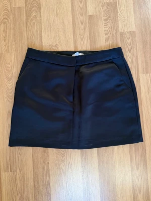 Svart kortkjol i satin-look H&M - Stilren svart kortkjol med lätt glansig satin-look och diskreta fickor framtill. Kjolen har en rak passform och bred linning som ger en clean och modern vibe. Den har också fickor. Perfekt att styla med en crop top eller oversized tröja.