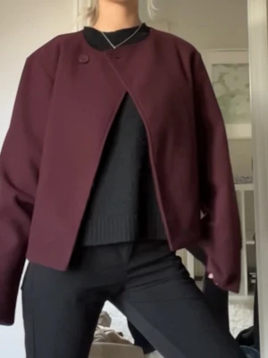 Vinröd kappa - Snygg vinröd kappa med omlott-design och en diskret knapp framtill. Jackan har en croppad passform och långa ärmar, perfekt för dig som vill ha en stilren och modern look. Materialet känns som ullblandning och färgen är mörk vinröd.