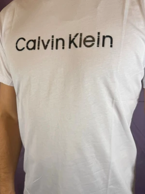 Vit Calvin Klein t-shirt med logga - Vit t-shirt från Calvin Klein med klassisk svart logga tryckt på bröstet. T-shirten har korta ärmar och en rak passform. Materialet är mjukt och känns som bomull, perfekt för en clean och stilren look.