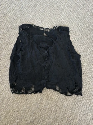 Svart spetslinnetopp med knappar - Snygg svart linnetopp med spetsdetaljer och knäppning framtill. Toppen har en croppad passform och fina broderade kanter. Perfekt att styla med höga jeans eller kjol för en trendig look.
