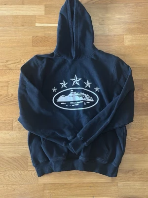 Corteiz Hoodie - Svart hoodie med stor vit grafik på bröstet som föreställer ett fartyg och fem stjärnor ovanför. Klassisk passform med huva och känguruficka. Mjuk insida och ribbade muddar. Perfekt för dig som gillar streetwear och snygga tryck.