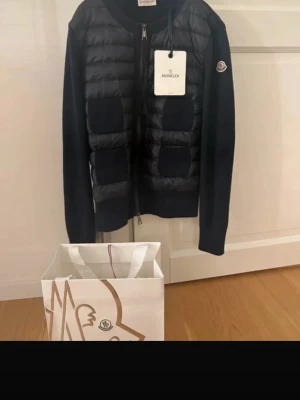 Marinblå cardiganjacka från Moncler - Snygg marinblå cardiganjacka från Moncler med quiltad front och fyra dekorativa fickor. Jackan har dragkedja framtill, ribbade muddar och är tillverkad i en mix av dun och stickat material. Moncler-logga på ärmen ger en exklusiv touch. POSTAR DAGEN DU KÖPER!