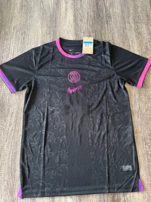 PSG Noir D.Doué #14 - Stilren svart fotbollströja med lila/rosa detaljer samt tryck på ryggen med D. Doué 14. Modern och unik design som passar perfekt för både samling, träning eller vardagsstil. Oanvänd!!! Fraktar dagen efter!!!