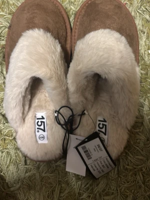 Bruna fluffiga tofflor från 157 - Mysiga bruna tofflor från 157 med supermjuk, ljusbeige fuskpäls på insidan och ovansidan. Yttermaterialet är mockaliknande och sulan är i slitstark gummi. Perfekta för att hålla fötterna varma hemma.