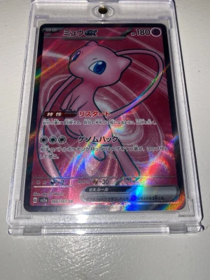 Mew GX Pokémonkort japansk holo - Säljer ett japanskt Mew GX Pokémonkort med holografisk effekt. Kortet har en rosa bakgrund med Mew i centrum och är inneslutet i ett genomskinligt plastfodral. Texten på kortet är på japanska och det har färgstarka detaljer.