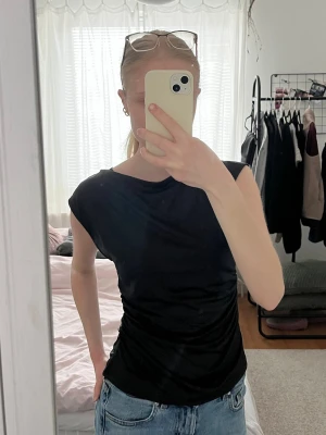 Svart topp från H&M med drapering - Säljer denna svarta topp från H&M med snygg drapering på sidan och breda axlar. Tunn och mjuk kvalitet, sidenliknande. Väldigt skönt material nu när det blir lite varmare! 