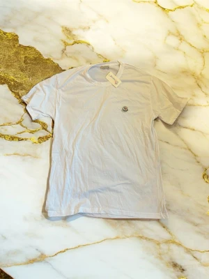 Vit t-shirt från Moncler - Vit t-shirt från Moncler med klassisk rund hals och korta ärmar. På bröstet finns en liten broderad Moncler-logga. T-shirten är gjord i mjuk bomull och har en clean, enkel look som funkar till allt. Perfekt för dig som gillar stilrena plagg med exklusiv känsla.