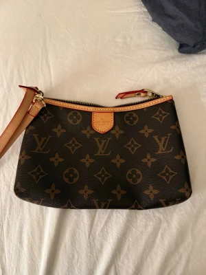 Clutch - Säljer en klassisk clutch i brunt monogrammönster med beige och röda detaljer. Väskan har guldfärgad dragkedja och avtagbar handledsrem i läder. Perfekt storlek. En liten defekt som syns på sista bilden men ingeting som man tänker på.