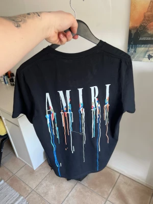 Svart AMIRI t-shirt med färgstänk - Säljer en svart t-shirt från AMIRI med stort tryck på ryggen och mindre tryck fram. Båda trycken har vita bokstäver och färgstänk i blått, rött och orangegga toner som rinner ner. Klassisk passform och rund hals. Perfekt för dig som gillar streetwear vibes.
