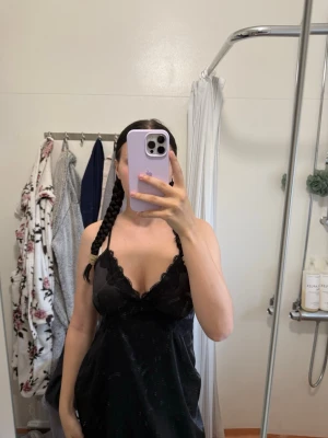 Svart spetsklänning med v-ringning - Svart klänning med v-ringad spetsdetalj och axelbandslös design. Klänningen har en kort längd och är tillverkad i ett mjukt syntetmaterial som ger en skön känsla. Perfekt för dig som gillar en stilren och elegant look.