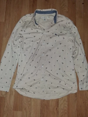 Beige skjorta med palmmönster - Snygg beige skjorta från Jack & Jones med små svarta palmer över hela tyget. Skjortan har långa ärmar, klassisk krage och knäppning framtill. Perfekt för en avslappnad och trendig look.