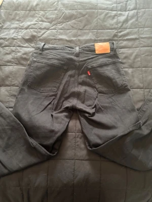 Svarta Levi's 501 jeans - Säljer ett par klassiska svarta Levi's 501 jeans med raka ben och fem fickor. Jeansen har den ikoniska läderpatchen bak i midjan och röd Levi's-tagg på bakfickan. Tillverkade i slitstark denim, perfekta för en tidlös look.