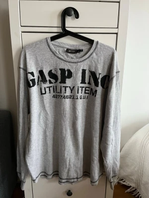 Grå långärmad tröja från GASP INC - Grå långärmad tröja från GASP INC med svart tryck framtill och baktill. Tröjan har rund halsringning, ribbade muddar och synliga sömmar. Tillverkad i ett mjukt, ribbat material som ger en avslappnad streetwear-look.