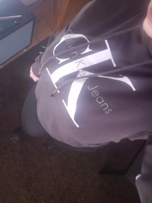 Svart hoodie från Calvin Klein Jeans - Säljer en svart hoodie från Calvin Klein Jeans med stor vit logga på bröstet och justerbar dragsko i huvan. Hoodien har en klassisk passform och är tillverkad i mjukt bomullsmaterial. Perfekt för en avslappnad och trendig look.