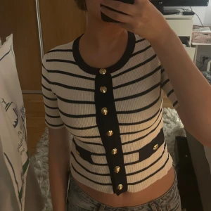 Randig ribbad topp med guldknappar - Säljer en kortärmad, ribbad topp med marinblå och vita ränder. Toppen har dekorativa guldknappar framtill, rund halsringning och två fejkfickor med knappdetaljer. Sitter tight och har en croppad passform. Perfekt för en trendig och chic look.