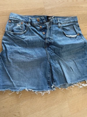 Blå jeansshorts med rå kant - Säljer ett par blå jeansshorts från IST med hög midja och rå, fransig kant nedtill. Klassisk femficksmodell med knappar i gylfen och slitna detaljer för en avslappnad look.