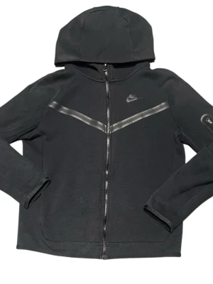 Svart Nike hoodie med dragkedja - Snygg svart hoodie från Nike med hel dragkedja framtill och huva. Har diskret Nike-logga på bröstet och en cool detalj med svarta ränder över bröstet. Tillverkad i mjukt material som känns skönt mot huden. Perfekt för en sportig och avslappnad stil.