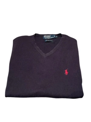 Mörklila tröja från Polo Ralph Lauren - Klassisk mörkblå tröja från Polo Ralph Lauren med V-ringning och den ikoniska röda logon broderad på bröstet. Tröjan har långa ärmar och ribbade muddar. Perfekt för en stilren och avslappnad look.