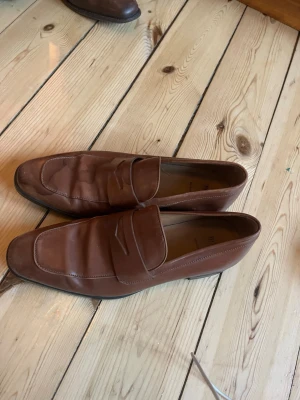 Bruna loafers från Hugo Boss - Snygga bruna loafers från Hugo Boss i äkta läder. Klassisk penny loafer-design med slät ovansida och diskret söm längs kanten. Tillverkade i Italien och har en elegant, tidlös look som passar till många stilar. Nypris 2200 kör 