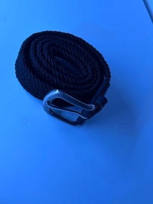 Svart flätat bälte med silverfärgat spänne - Snyggt svart flätat bälte med ett stilrent silverfärgat metallspänne. Bältet har en klassisk design och passar perfekt till jeans eller chinos. Gjort i syntetmaterial med detaljer i skinnimitation.