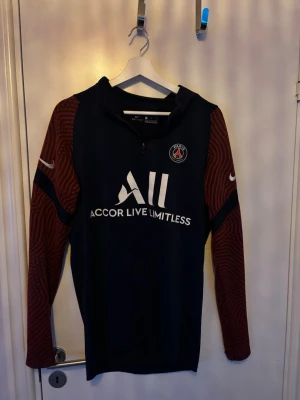 PSG träningsjacka Nike blå/röd - Säljer en PSG träningsjacka från Nike i mörkblått med röda mönstrade ärmar. Jackan har halv dragkedja framtill, PSG-logga på bröstet och Nike-logga på ärmarna. Materialet är syntetiskt och passar perfekt för sport och träning.