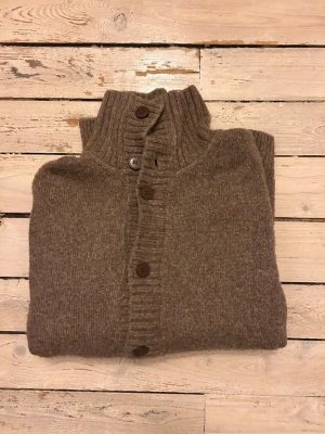 Brun stickad kofta med knappar - Mysig brun stickad kofta med ribbad krage och knäppning framtill. Koftan har en klassisk design med grovstickade detaljer längs knappraden och kragen. Perfekt för lager-på-lager och chill dagar.