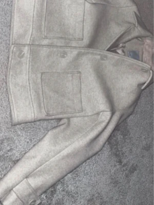 Beige cardiganjacka med fickor - Stilren beige cardiganjacka med raka linjer och två stora framfickor. Jackan har knäppning framtill och är tillverkad i ett mjukt, tjockare tyg som ger en clean look. Perfekt för lager-på-lager och enkel att matcha.