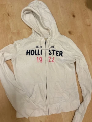 Vit hoodie från Hollister zip - Den klassiska hoodien från Hollister, med marinblå o rosa tryck.