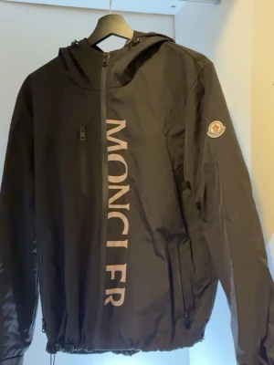 Windbreaker moncler  - Snygg svart vindjacka från Moncler med stor logga längs dragkedjan och klassisk Moncler-patch på ärmen. Jackan har huva, dragkedjeförsedda fickor och justerbar nederkant. Tillverkad i lätt och slitstarkt material, perfekt för blåsiga dagar.