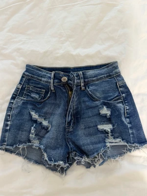 Blå högmidjade jeansshorts - Säljer ett par blå högmidjade jeansshorts med slitna detaljer och fransig kant. Shortsen har klassisk femficksdesign och dragkedja med knapp. Perfekta för sommaren och ger en avslappnad vibe.