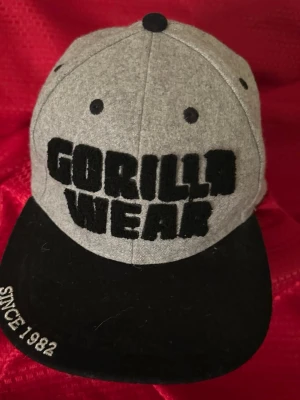 Grå och svart keps Gorilla Wear - Snygg keps från Gorilla Wear i grått med svart skärm och broderad text framtill. Skärmen är platt och framtill står det 'GORILLA WEAR' i stora bokstäver samt 'SINCE 1982' broderat på sidan av skärmen. Perfekt för en streetwear-look.