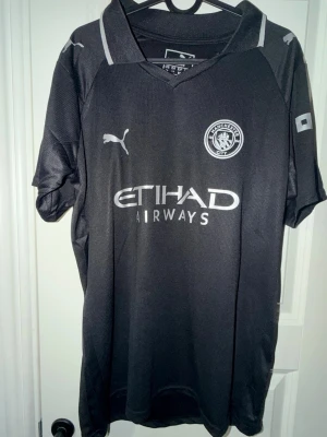 Manchester City svart matchtröja Puma - Svart Manchester City matchtröja från Puma med klubbmärke och Etihad Airways-logga på bröstet. Tröjan har korta ärmar, sportig passform och är tillverkad i ett lätt, ventilerande material perfekt för fotboll eller träning.